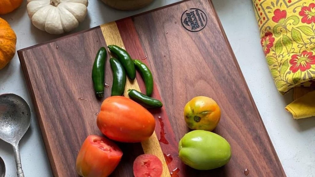 Koshur Chaan Woodworking Handmade hardwood cutting board with sliced tomatoes, green chilies, gourds, spoon, and floral cloth on kitchen counter. Johns Creek, GA