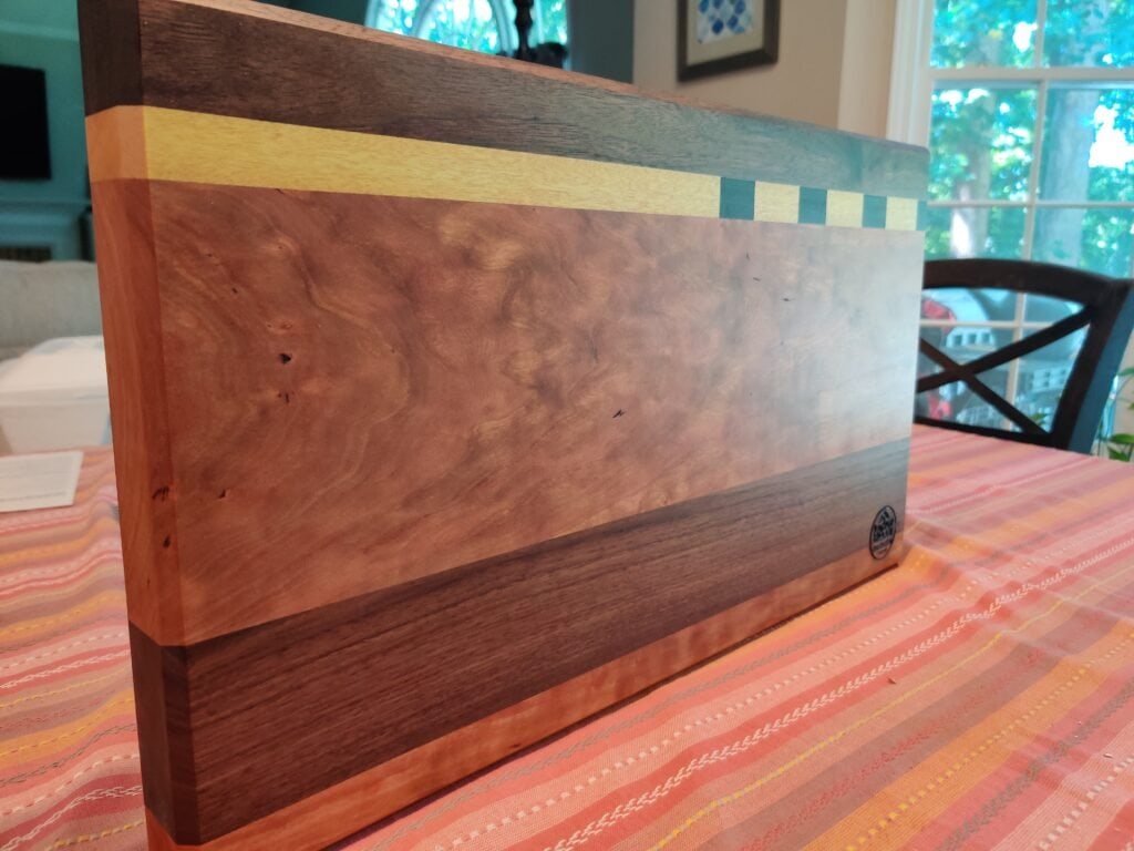 Koshur Chaan Woodworking Handmade hardwood cutting board with walnut, cherry, and maple accents, upright on a striped tablecloth. Artisan crafted in Georgia. Johns Creek, GA