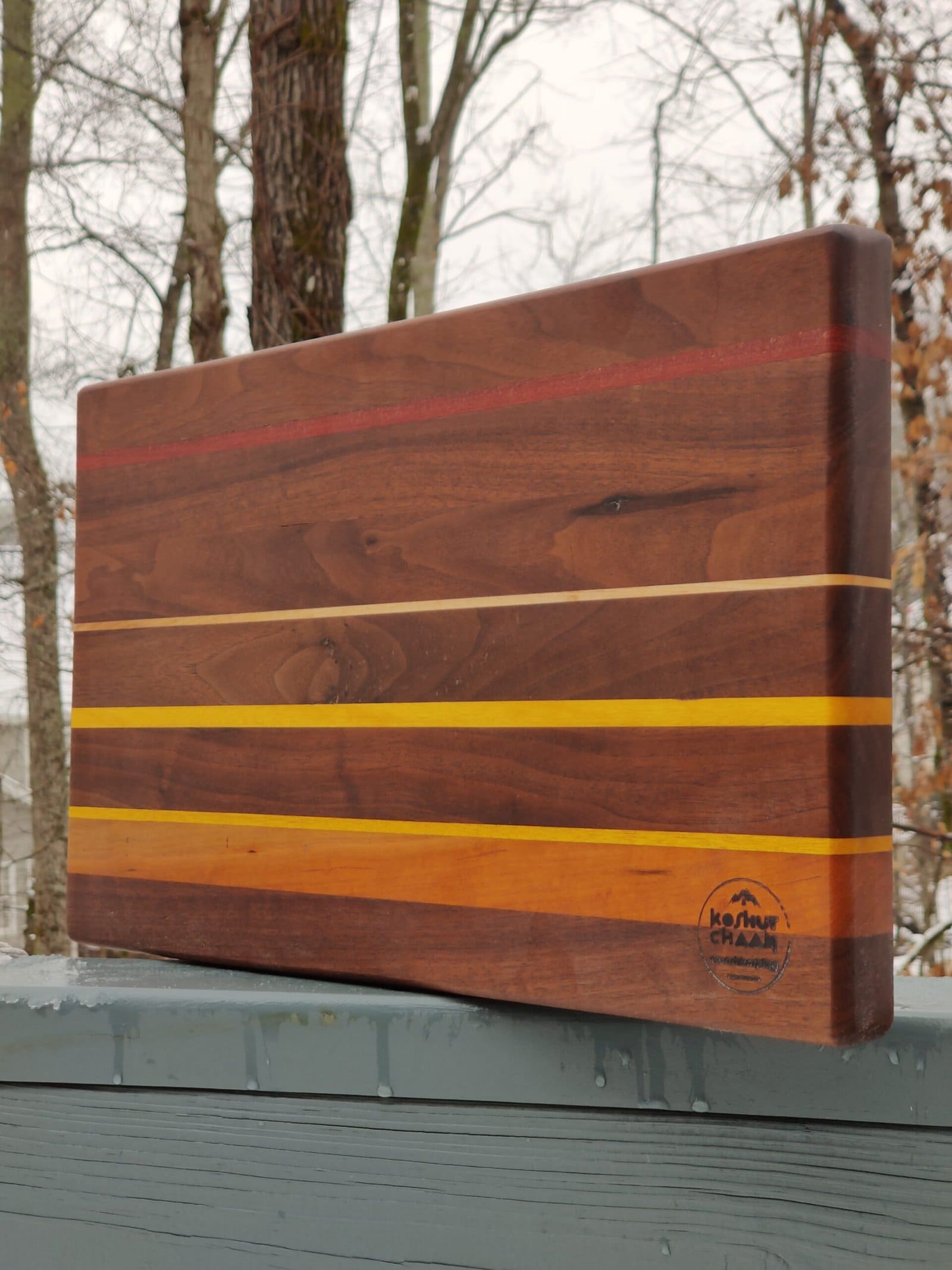 Koshur Chaan Woodworking Artisan handmade cutting board with maple, cherry, and walnut inlaid stripes; maker's logo; snowy Georgia background. Johns Creek, GA