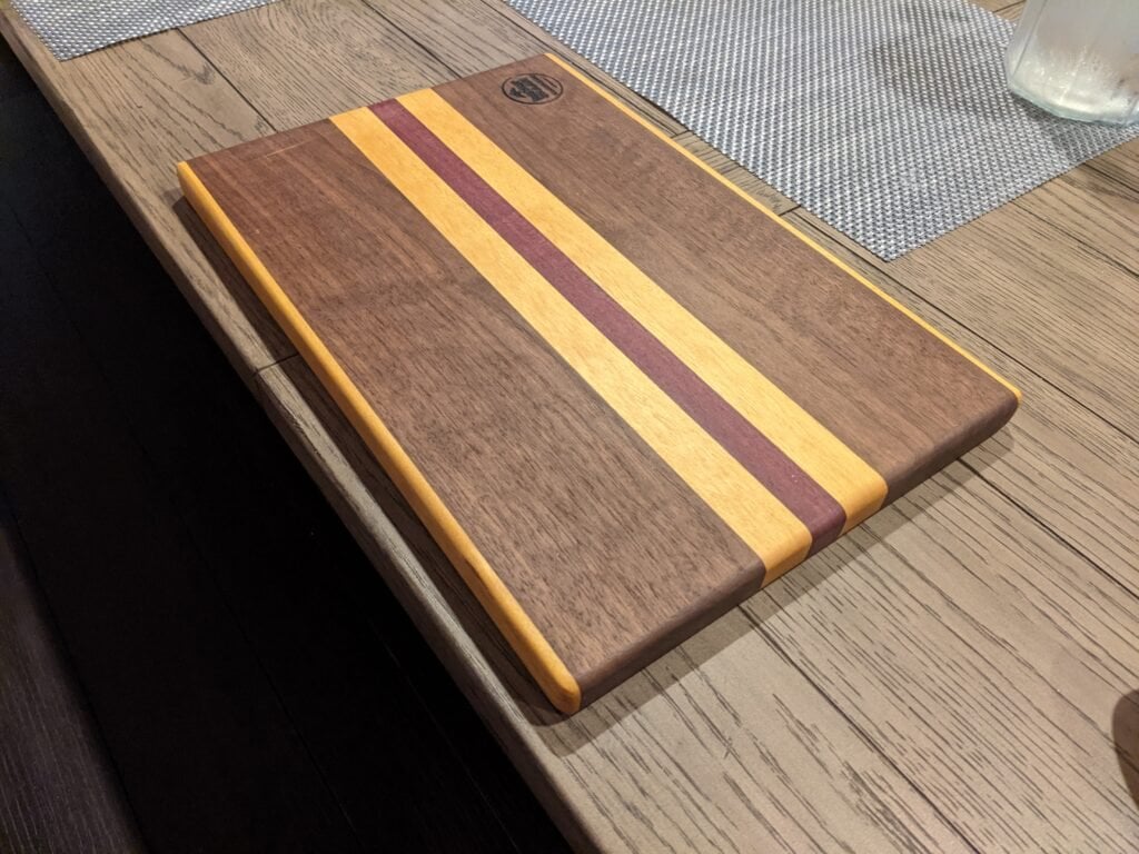 Koshur Chaan Woodworking Handmade hardwood cutting board with walnut, maple, and cherry stripes, engraved logo in corner, on a wooden table with placemats. Johns Creek, GA