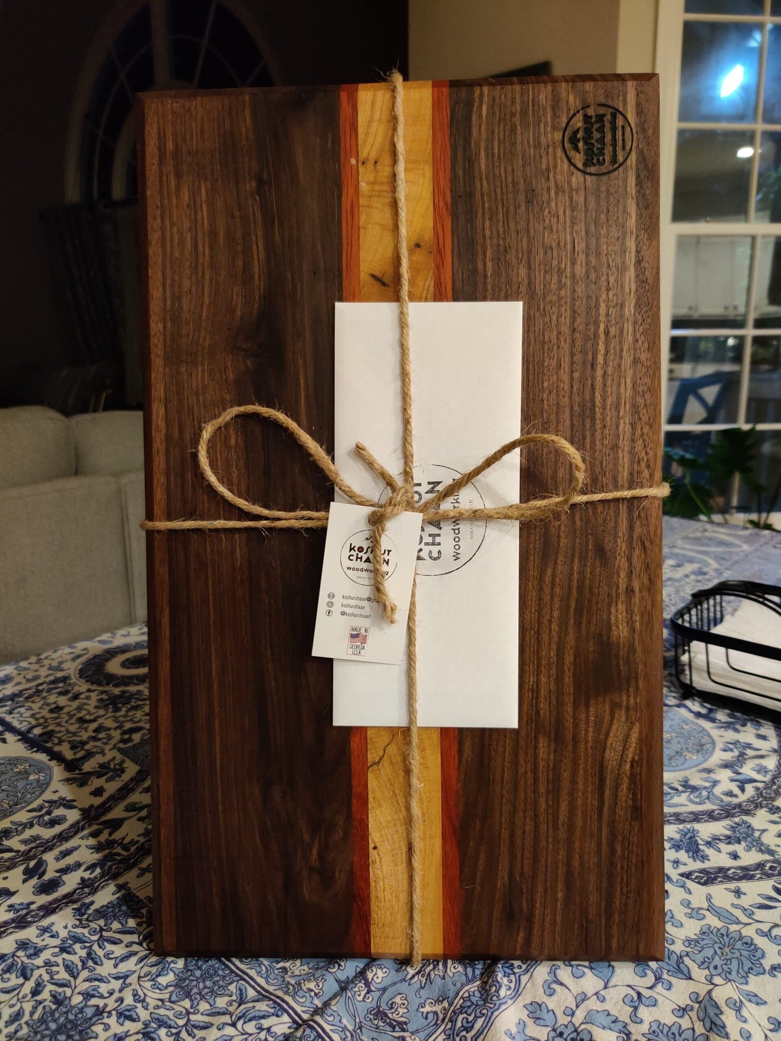 Koshur Chaan Woodworking Handmade artisan cutting board with walnut and maple stripes, logo stamp, and tied gift card on a blue patterned tablecloth. Johns Creek, GA