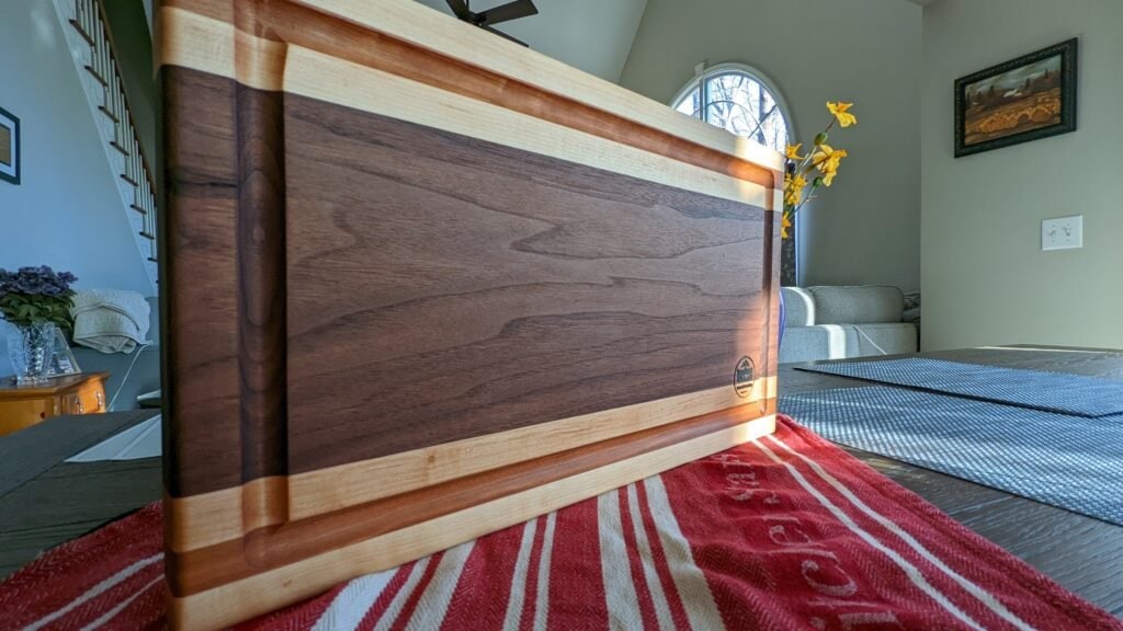 Koshur Chaan Woodworking Handmade hardwood cutting board with walnut and maple stripes on a dining table; sunlight highlights artisan craftsmanship. Johns Creek, GA