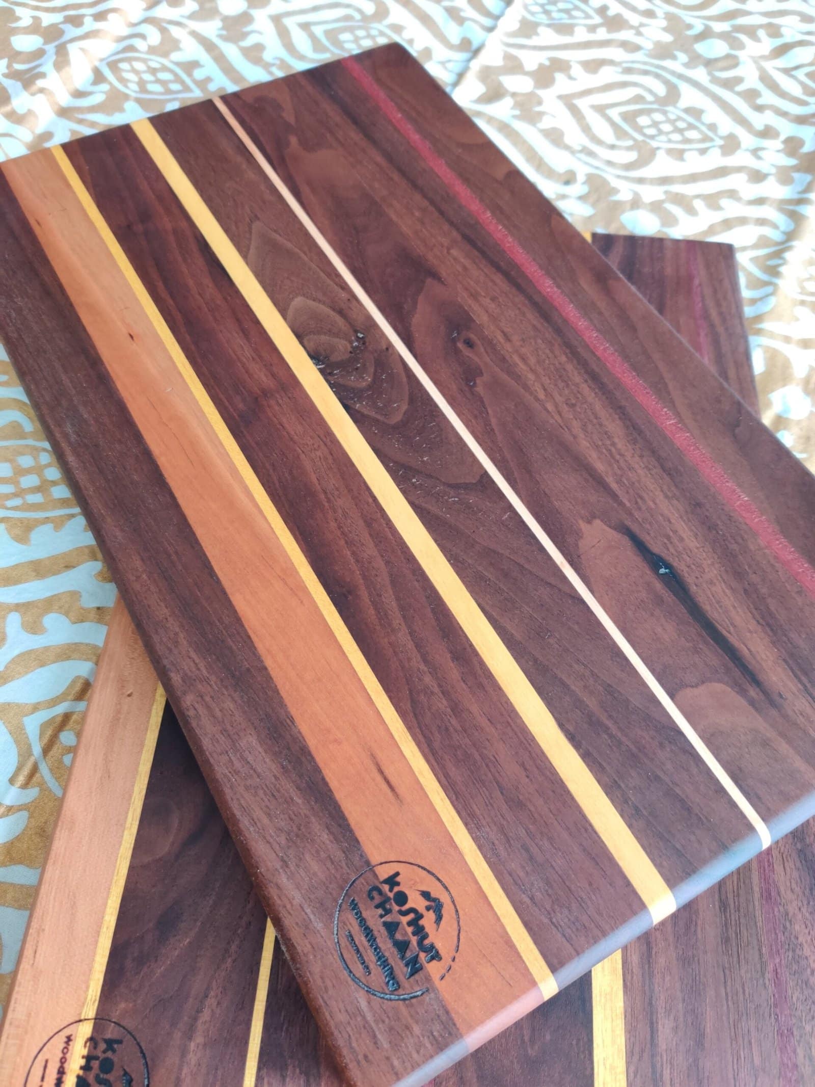 Koshur Chaan Woodworking Two artisan hardwood cutting boards with walnut, maple, and cherry stripes, stamped “Loamy Crown Creations,” on patterned fabric. Johns Creek, GA