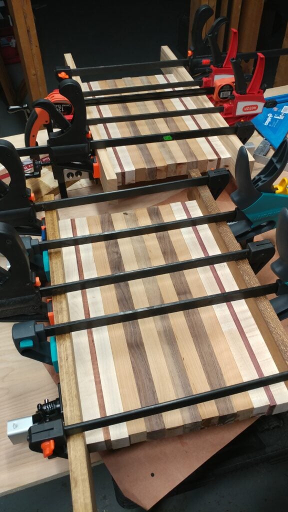 Striped cutting board assembly with visible end grain secured in black bar clamps with red and orange handles