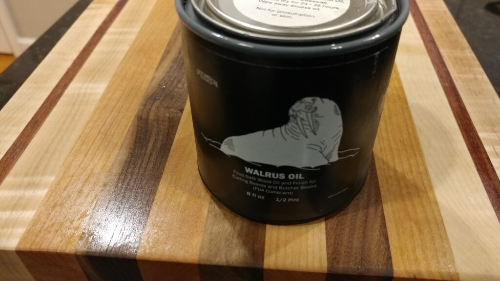 Striped wooden cutting board beside a dark container of board finish on a work surface