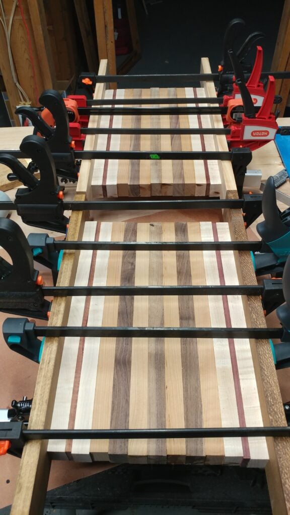 Striped wooden cutting board secured across a workbench by black bar clamps with red and orange handles