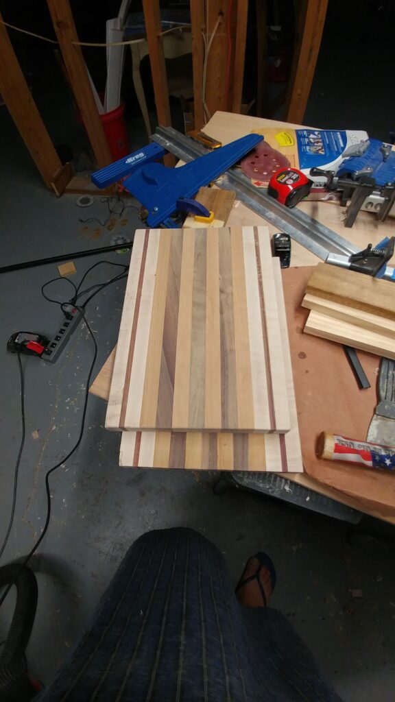 Part of the Koshur Chaan woodworking story: striped board pieces on a workbench with blue clamps, a tape measure and sanding discs
