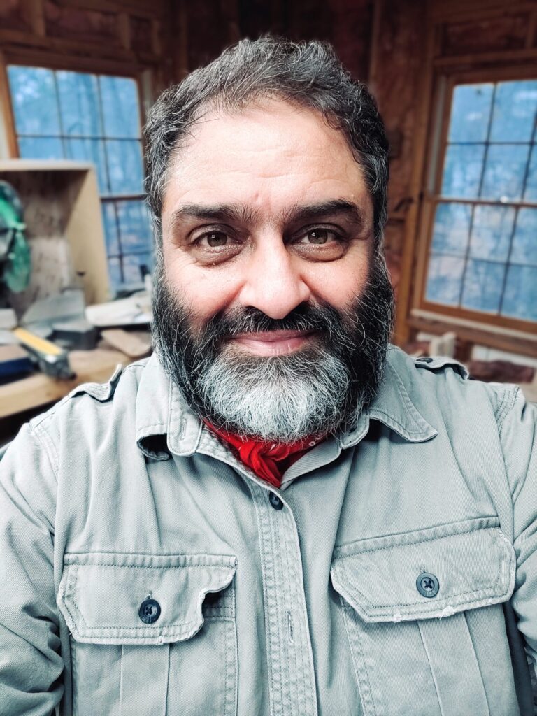 Ishtyaq Qadri, known as Wosta, smiling in his Johns Creek woodworking shop, wearing a gray work shirt and red bandana
