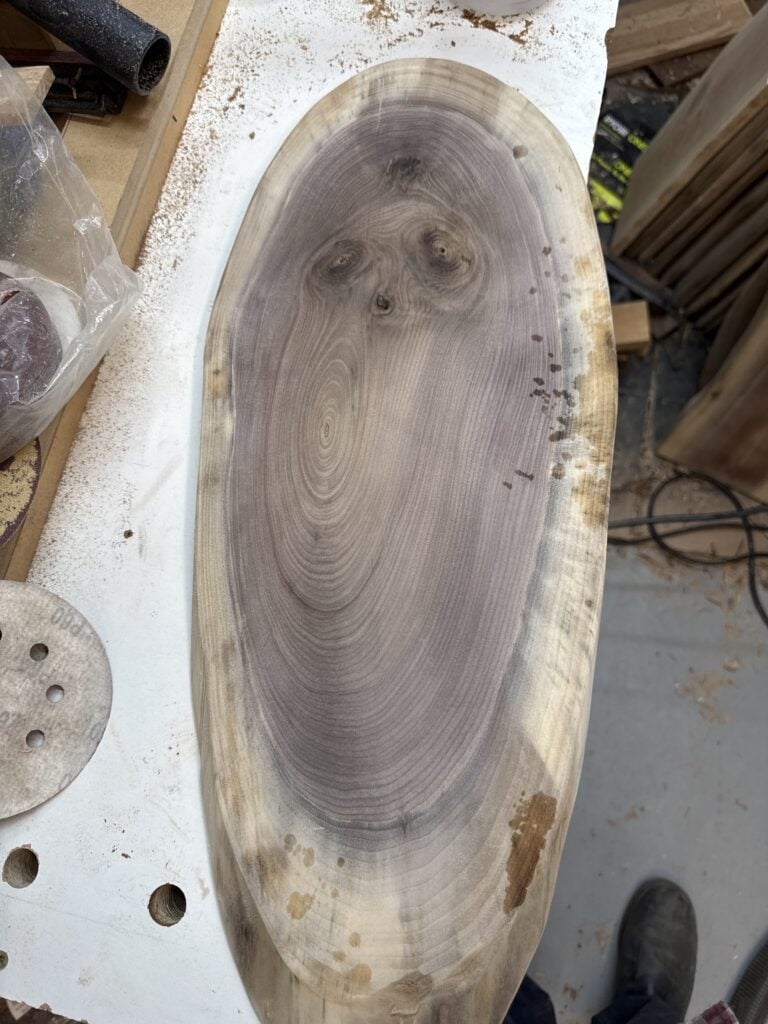 Oval wood slab with dark grain, knots and a pale natural edge on a workshop bench