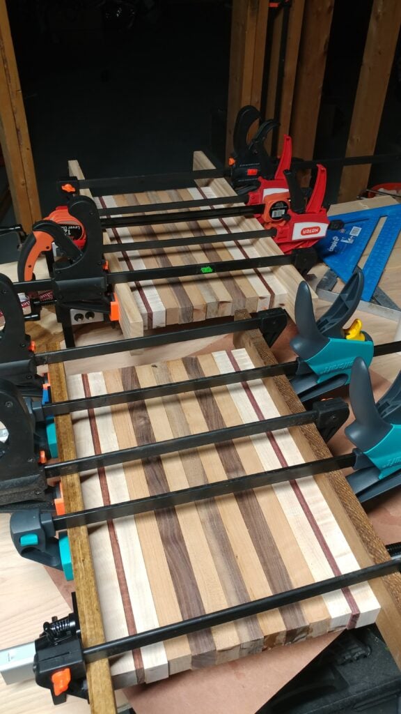 Striped wooden cutting board held with black, red and orange clamps on a workshop bench